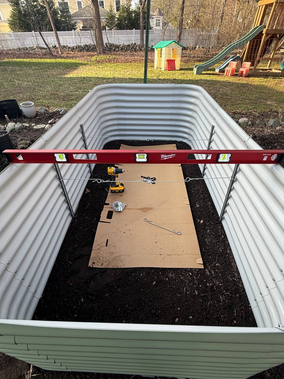 Interior of a large Birdies raised bed looking down, with a Milwaukee level across the top, a DeWalt drill, and cardboard laid over the soil